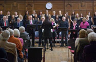 A choir singing in a hall, their conductor facing them, with an audience seated in front